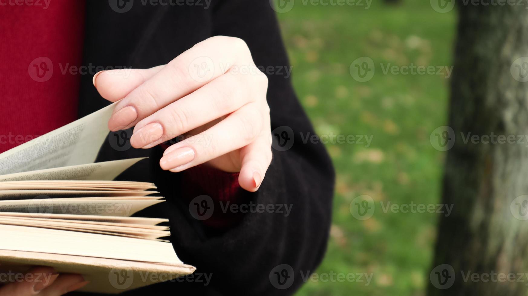 flicka läser en bok i parken. kvinnliga händer bläddra sidor av pappersbok utomhus. studenten förbereder sig för tentamen. litterär fritid i naturen. närbild, kopiera utrymme. foto