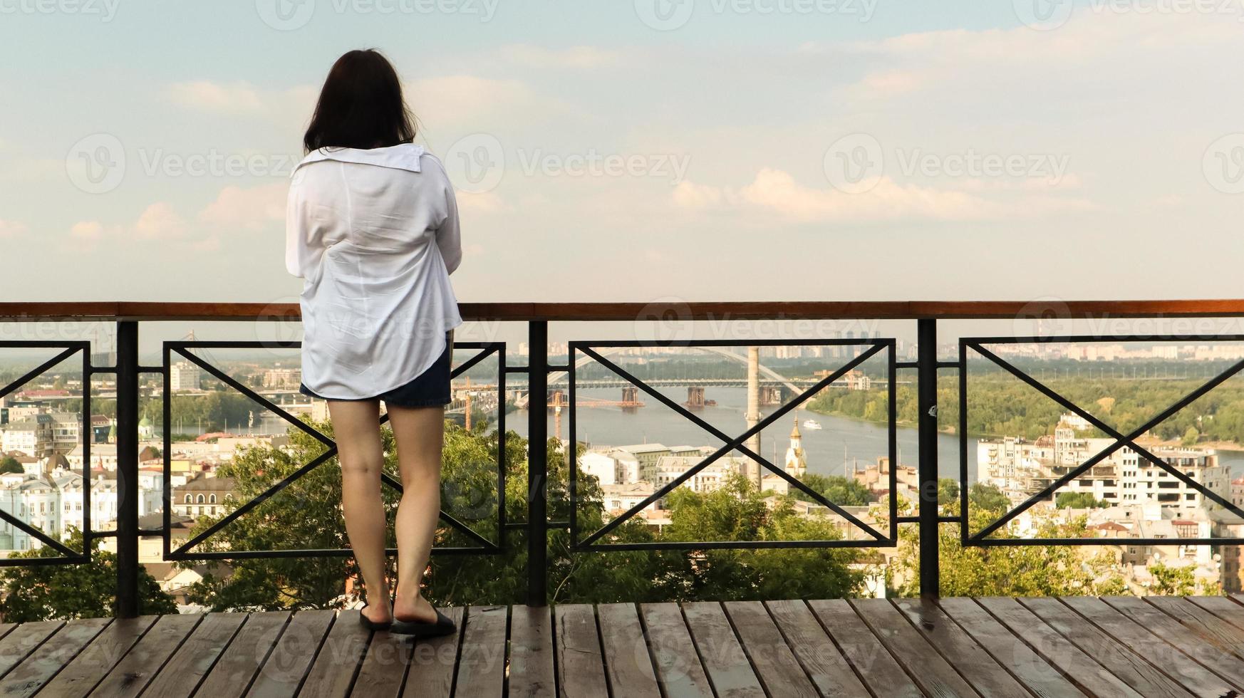 bakifrån av en ung kvinna i vit skjorta, en brunettresenär tittar på stadsbilden en solig dag från en hög kulle med ett observationsdäck för turister. årgång foto