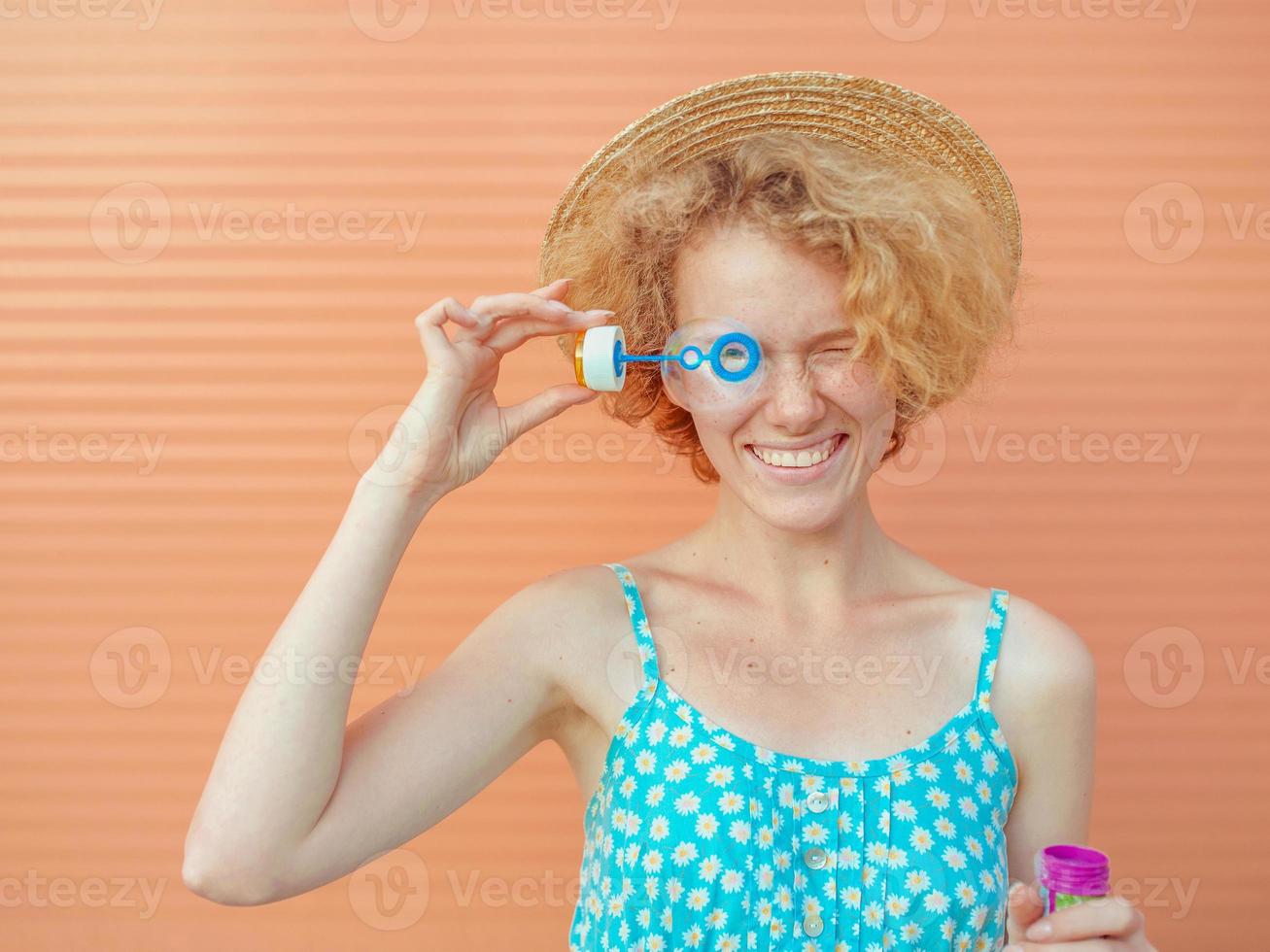 ung glad, lockig rödhårig kvinna i blå sundress och halmhatt med bubblor på beige bakgrund. kul, sommar, mode, ungdomskoncept foto