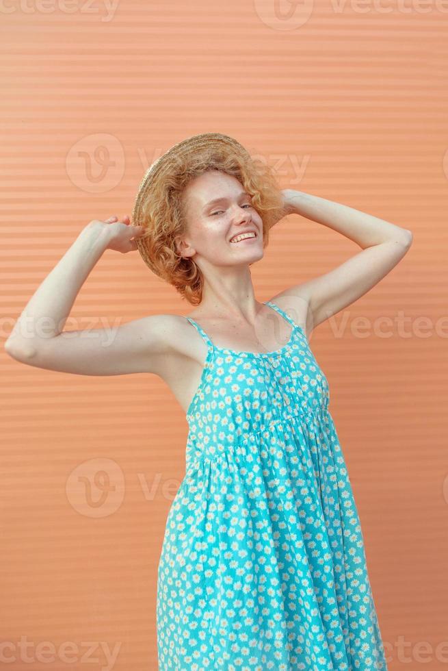 ung glad, lockig rödhårig kvinna i blå sundress håller stråhatt i handen på beige bakgrund. kul, sommar, mode, ungdomskoncept foto