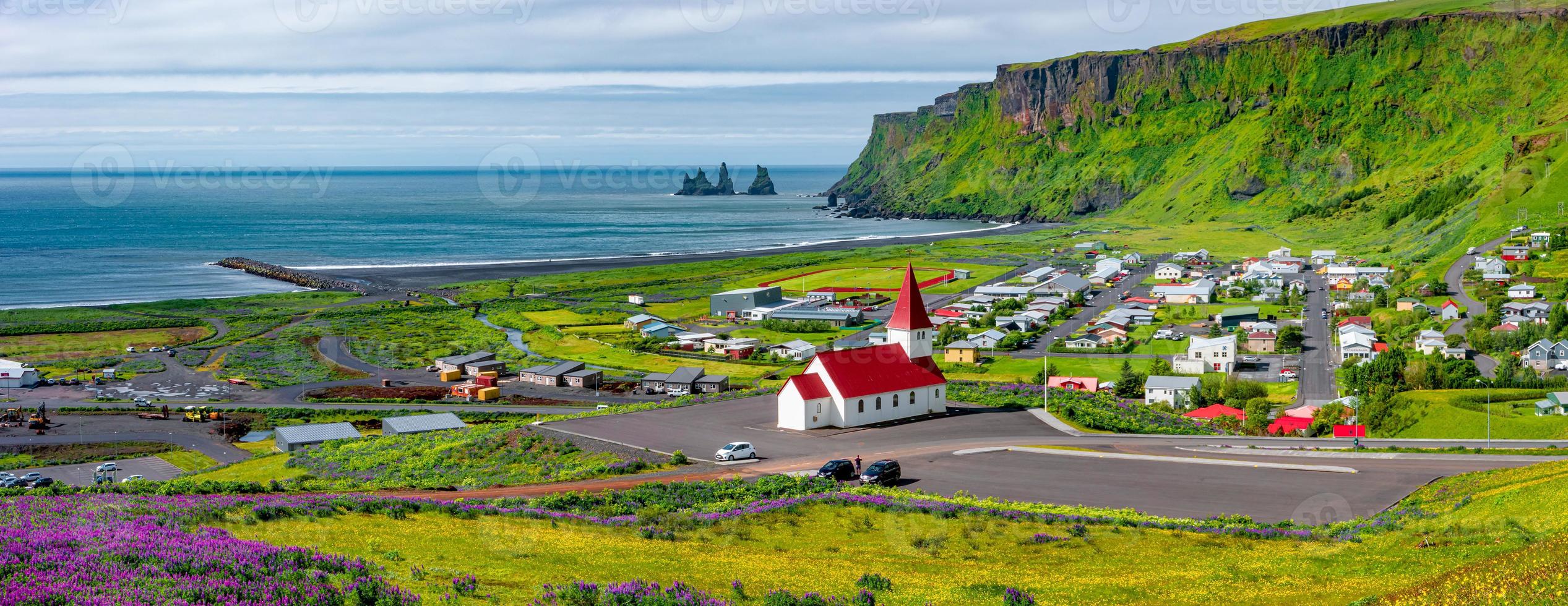 panoramautsikt över basaltstackar reynisdrangar, vulkanisk svart sandstrand och violetta lupin och gula ängsblommor vid vik stad, och en luthersk kyrka, södra island, vid solig sommardag blå himmel. foto