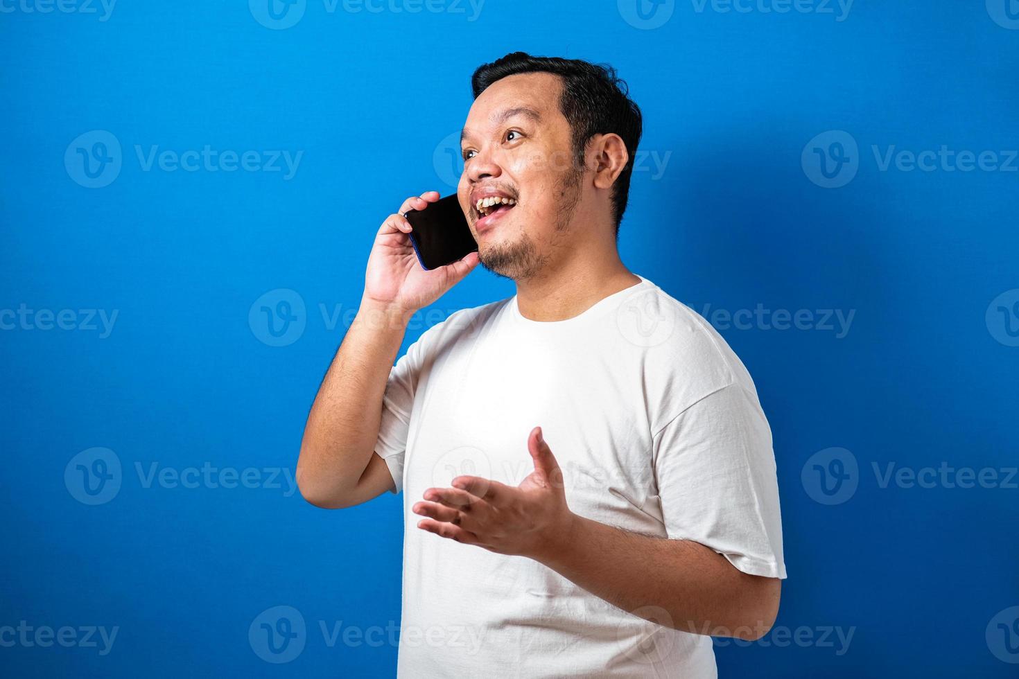 fet asiatisk man i vit t-shirt ser glad ut medan han pratar på smartphone. halva kroppen studio skott mot blå bakgrund foto