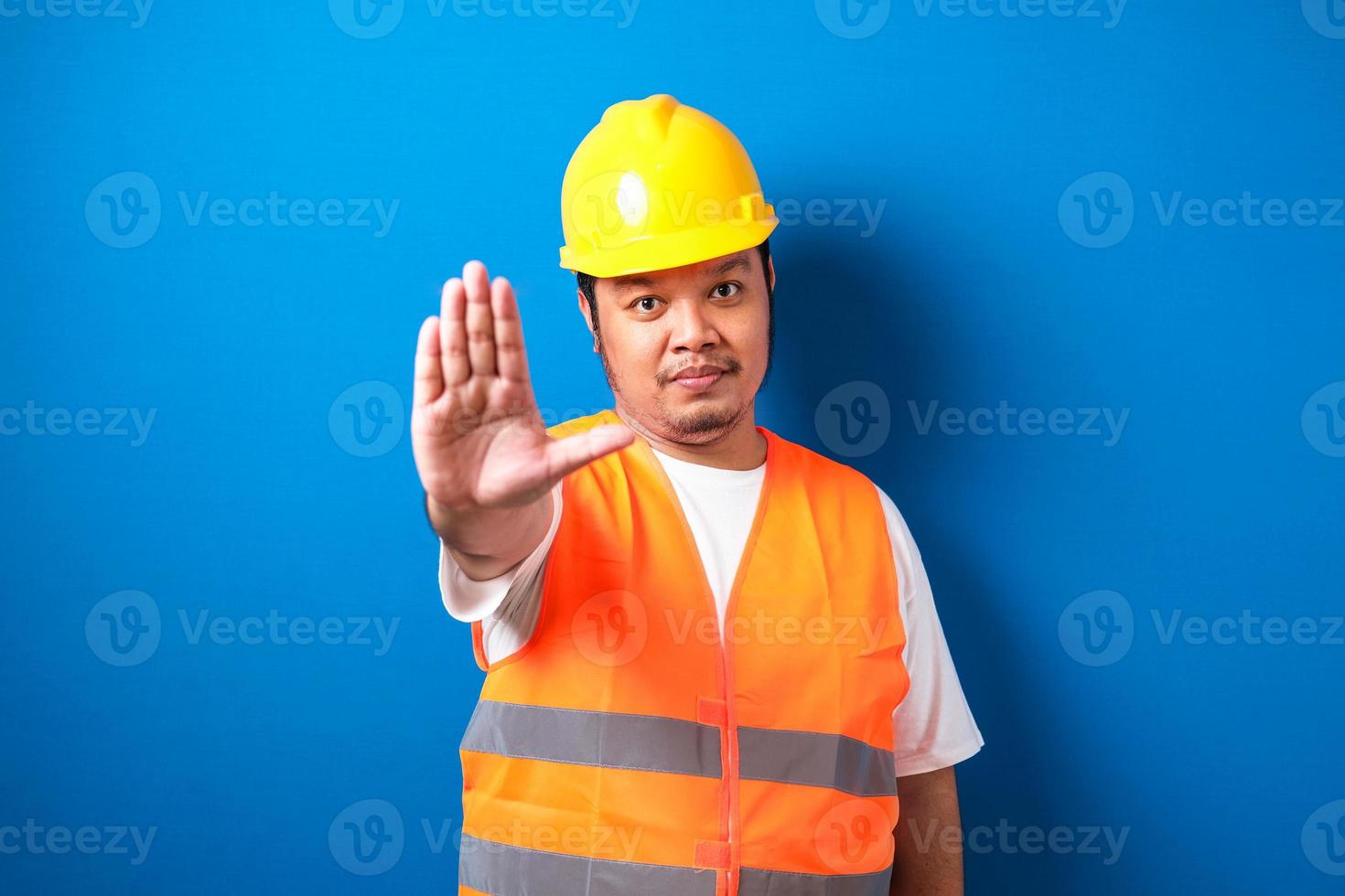 fet asiatisk arbetare som bär orange säkerhetsväst och gul hjälm gör stoppgest foto