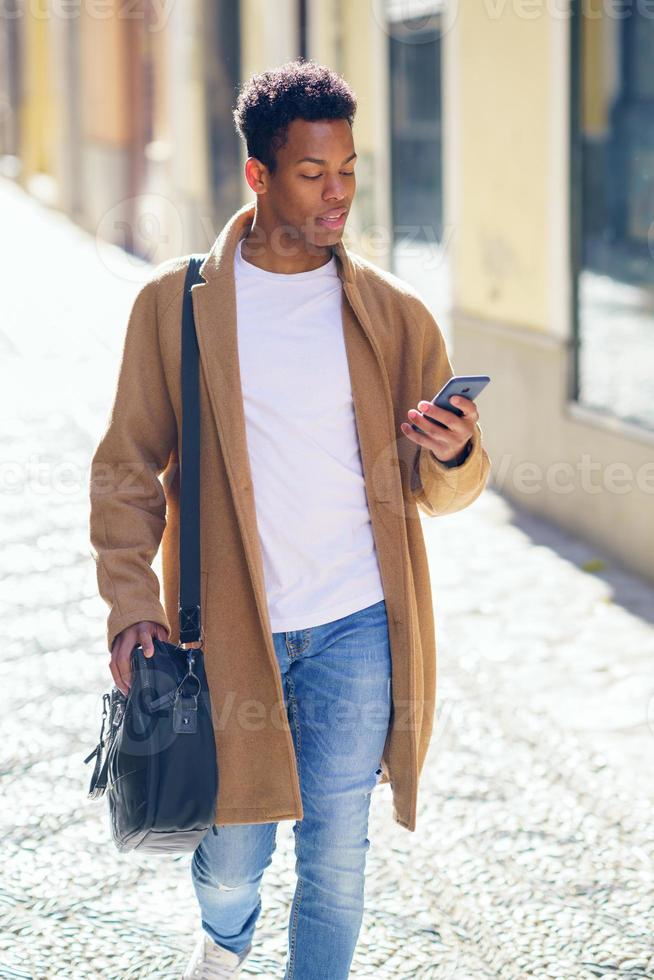 ung svart man går nerför gatan med en portfölj och en smartphone. foto