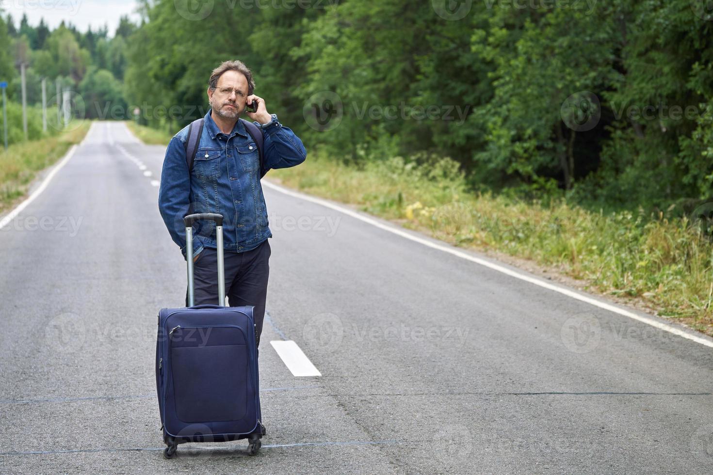 man med ett bagage som står mitt på en asfaltsväg och pratar i mobiltelefon foto