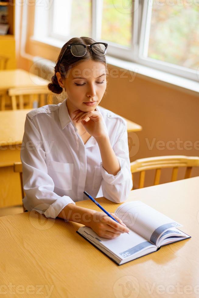 söt student sitter med en anteckningsbok vid skrivbordet vid fönstret, omtänksam utsikt foto