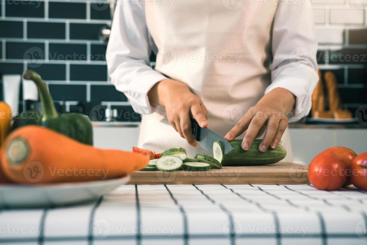 hemmafru med kniv och händer skär gurka på träskiva i köksrummet. foto