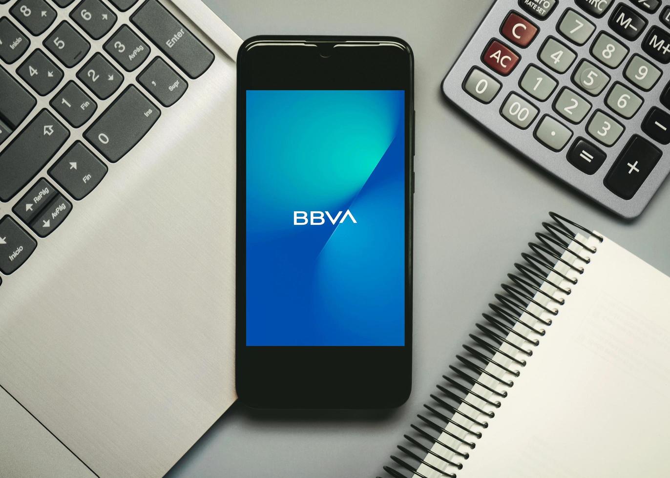 bbva mobilapps logotyp på smartphoneskärmen bredvid en anteckningsbok, bärbar dator och miniräknare foto