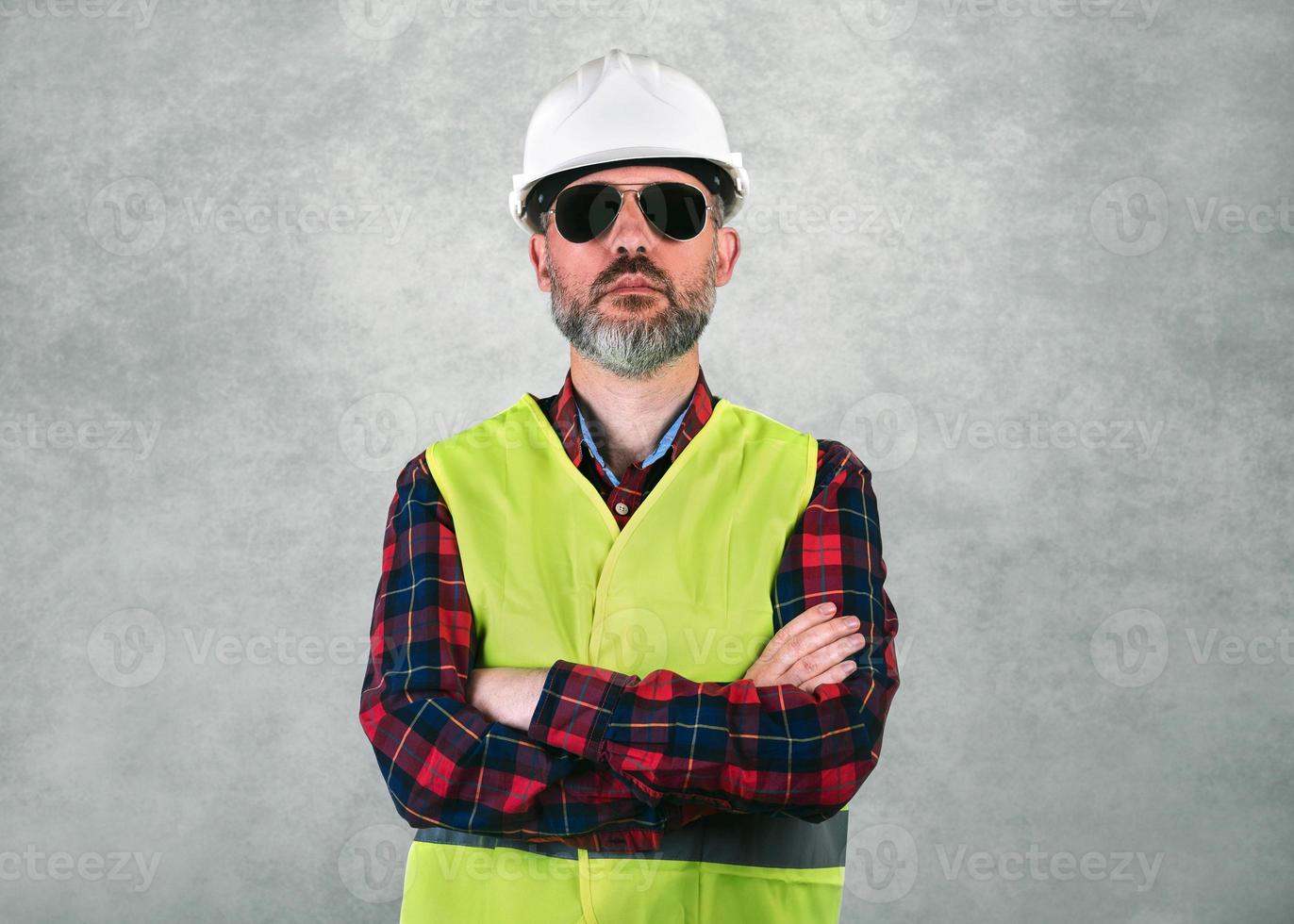 man byggnadsarbetare med en vit hjälm och solglasögon med armarna i kors och säkerhetsväst stående foto