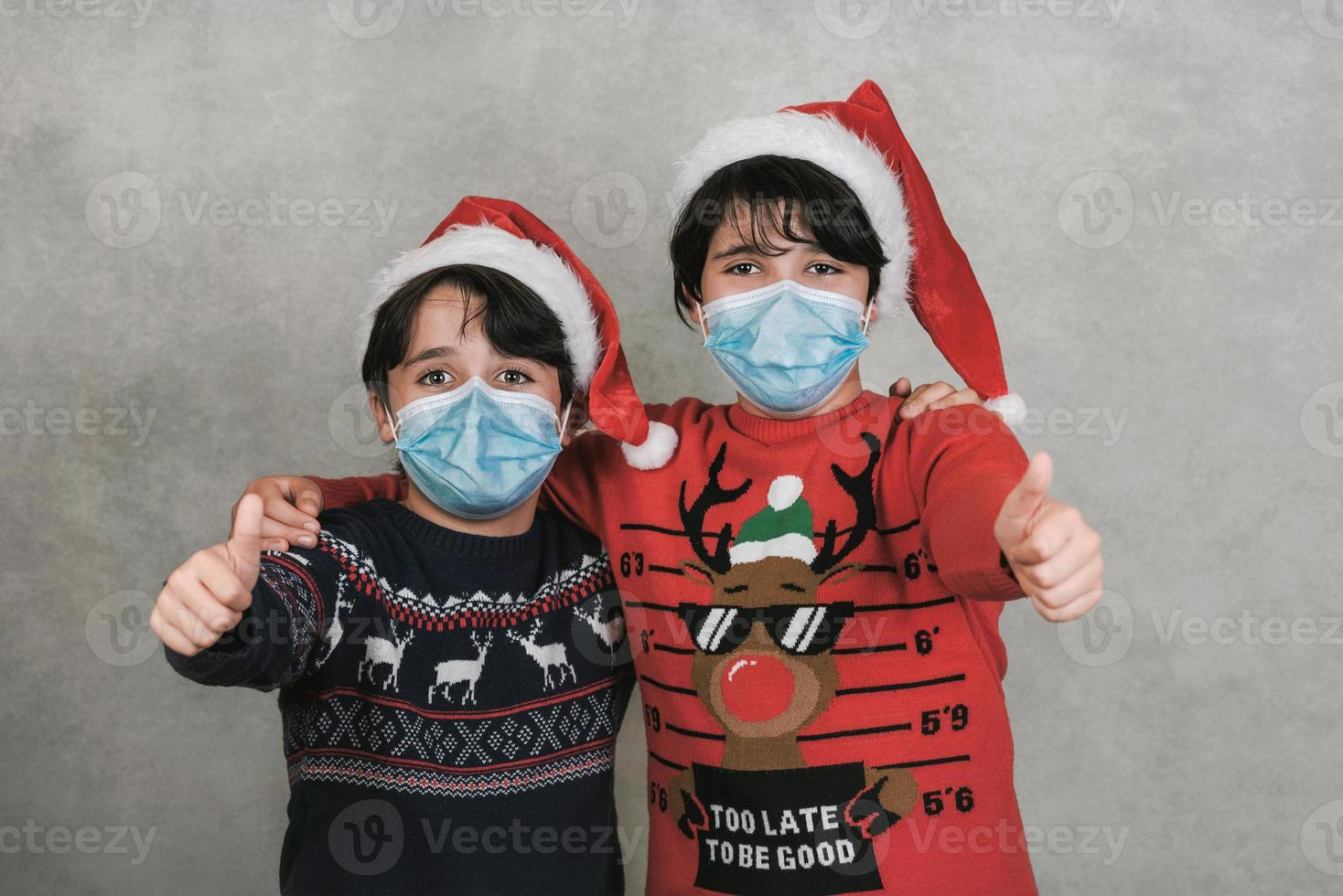 god jul, två barn med medicinsk mask och jultomtens hatt gör segertecken foto