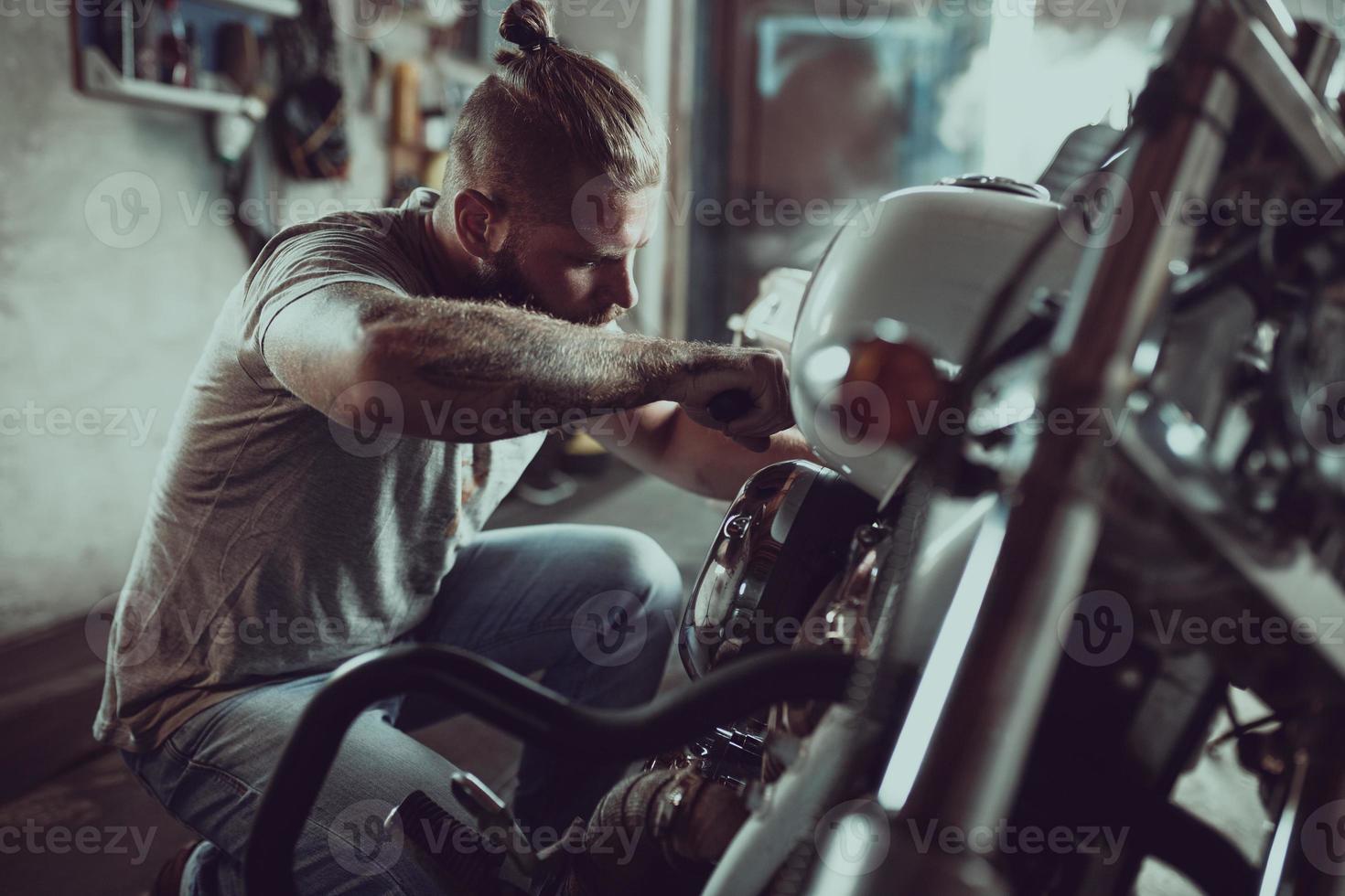 stilig skäggig man reparerar sin motorcykel i garaget. en man klädd i jeans och en t-shirt foto