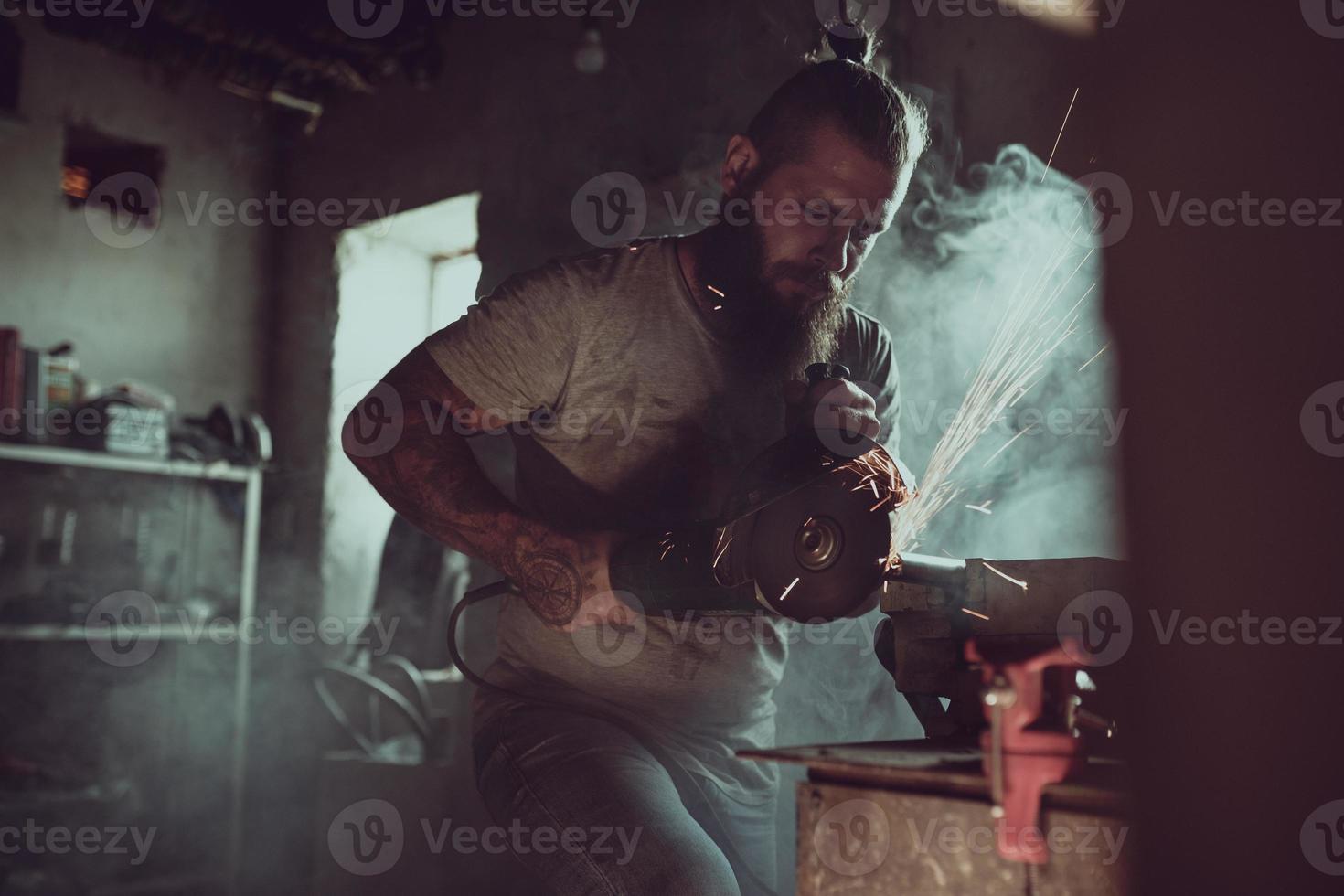 stilig brutal man med skägg som reparerar en motorcykel i sitt garage och arbetar med en cirkelsåg. i garaget mycket gnistor och rök från sågning foto