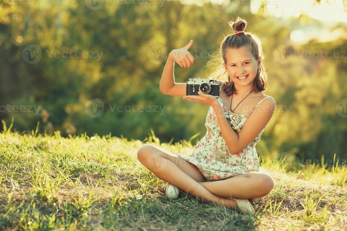 barn flicka sitter på gräset. i händerna har hon en gammal fotokamera och hon visar gester okej foto