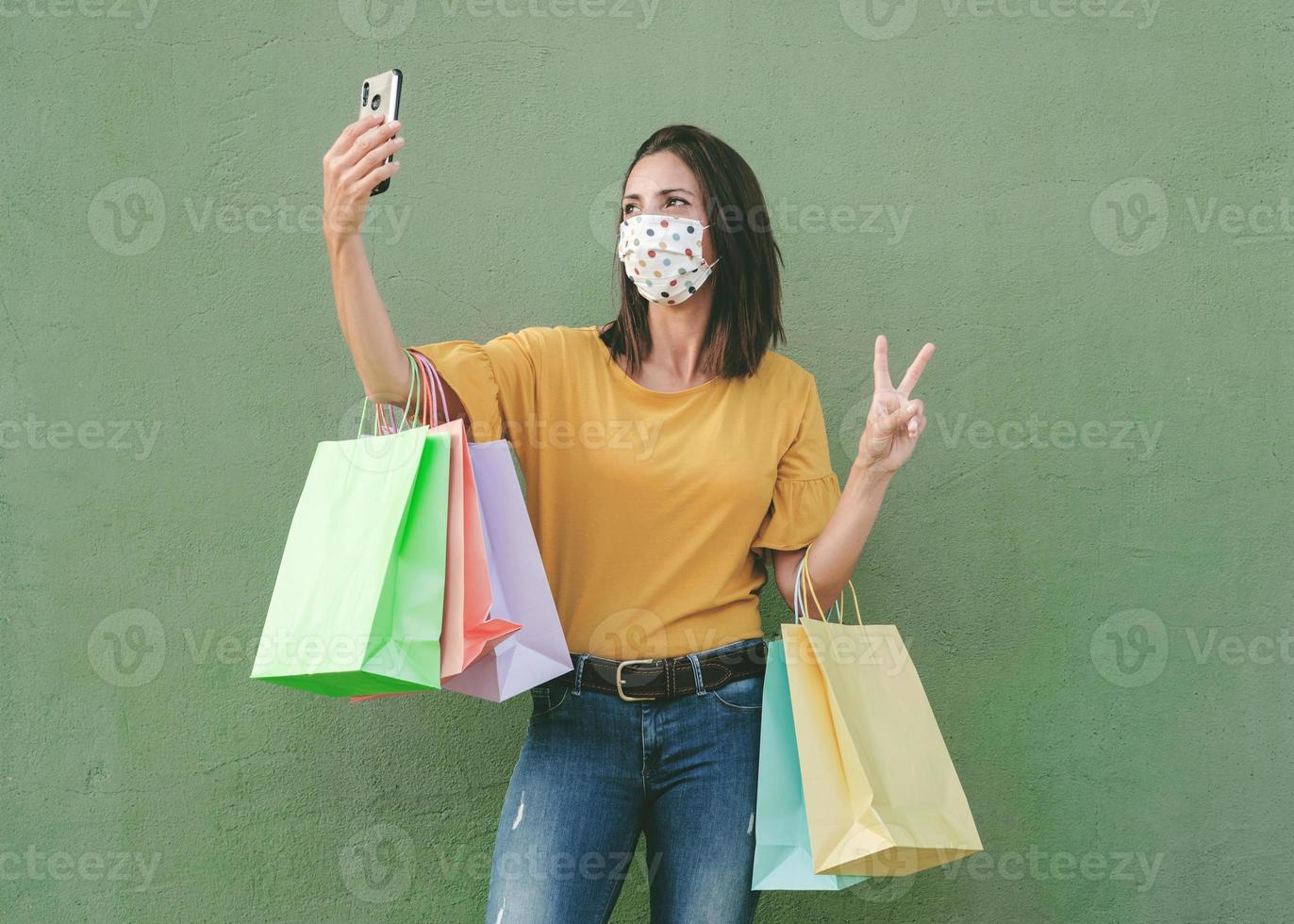 ung glad kvinna bär medicinsk mask som håller en shoppingpåsar tar en selfie på smartphone foto