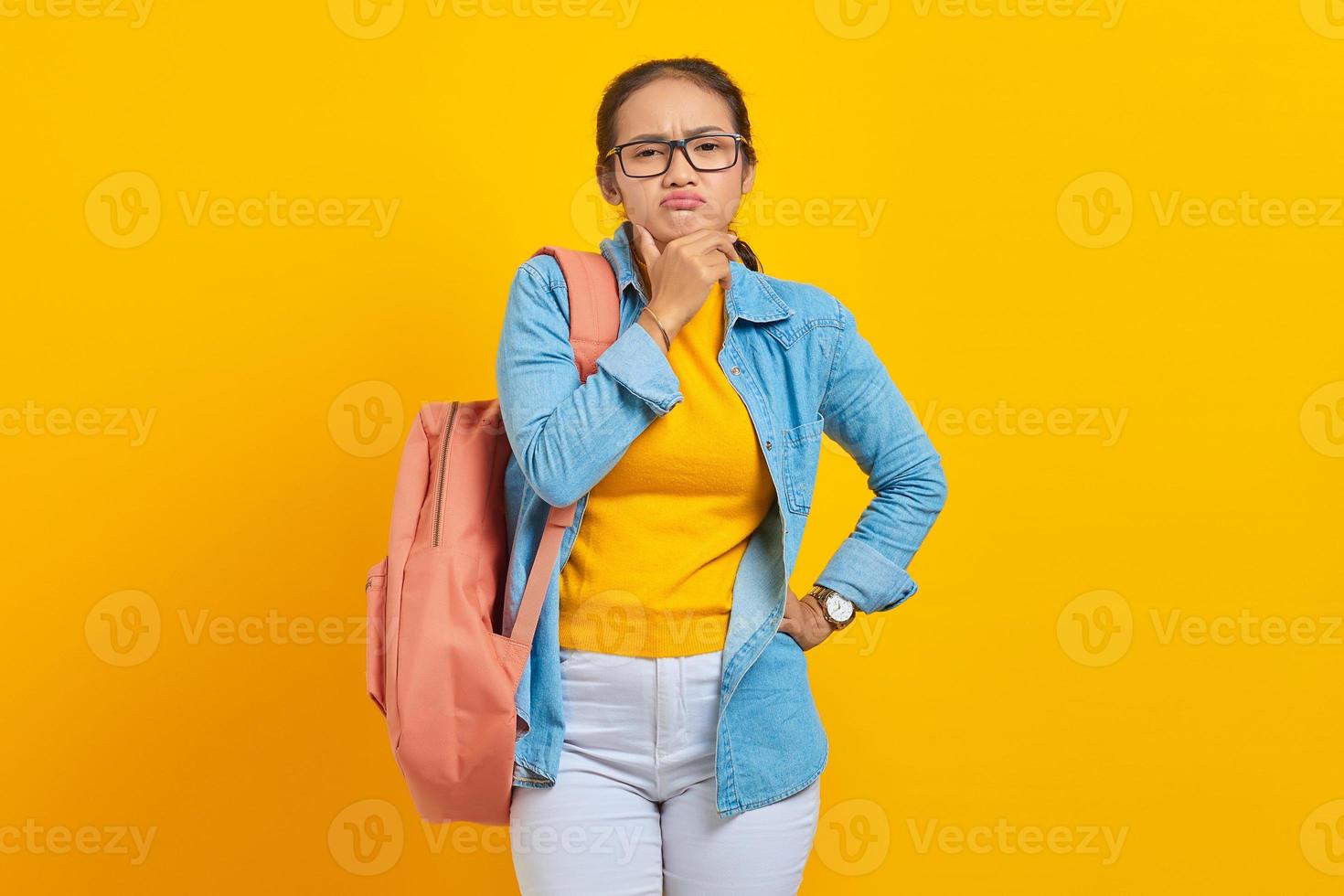 eftertänksam ung asiatisk student i denim outfit med ryggsäck håller hakan medan du tänker något isolerat på gul bakgrund. utbildning i gymnasiekonceptet foto