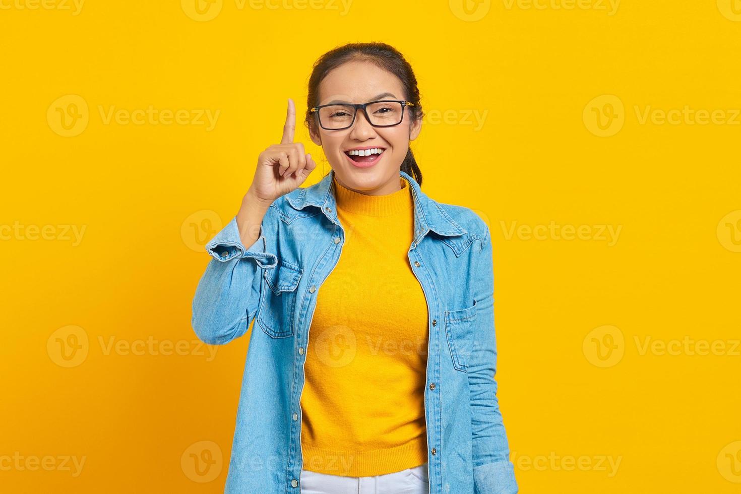 porträtt av glad ung asiatisk kvinna student i jeanskläder med kreativ idé och pekar finger uppåt isolerad på gul bakgrund. utbildning i college universitet koncept foto