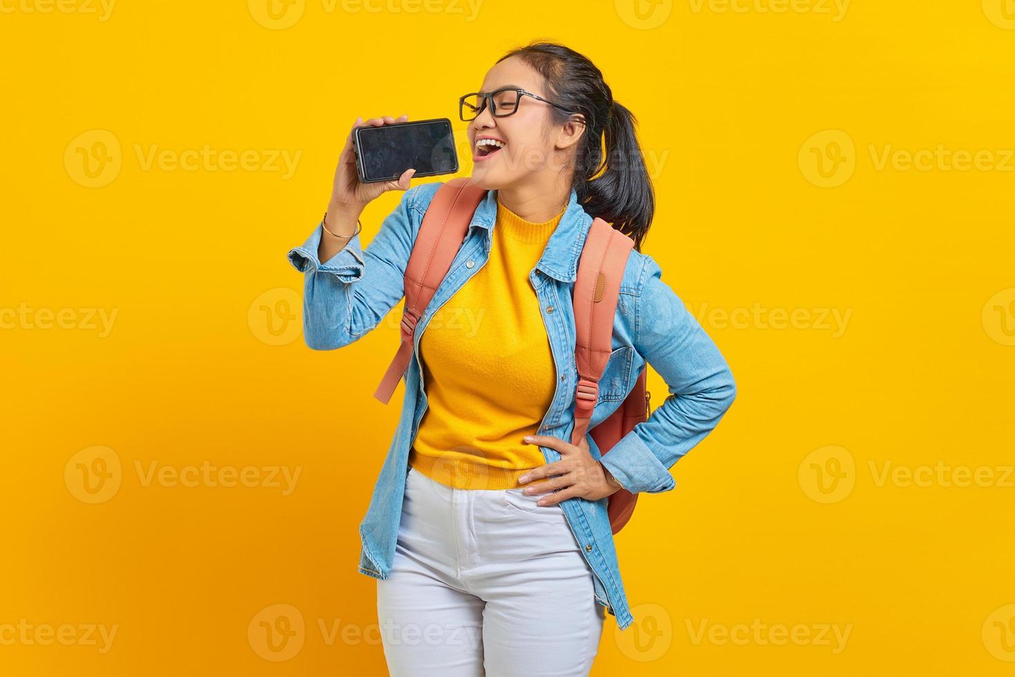 porträtt av leende ung asiatisk kvinna student i fritidskläder med ryggsäck njuter av att sjunga med smartphone isolerad på gul bakgrund. utbildning i college universitet koncept foto