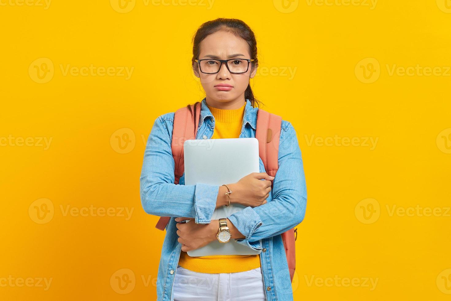 porträtt av missnöjd ung asiatisk kvinna student i fritidskläder med ryggsäck som håller laptop och tittar på kameran isolerad på gul bakgrund. utbildning i högskolekoncept foto