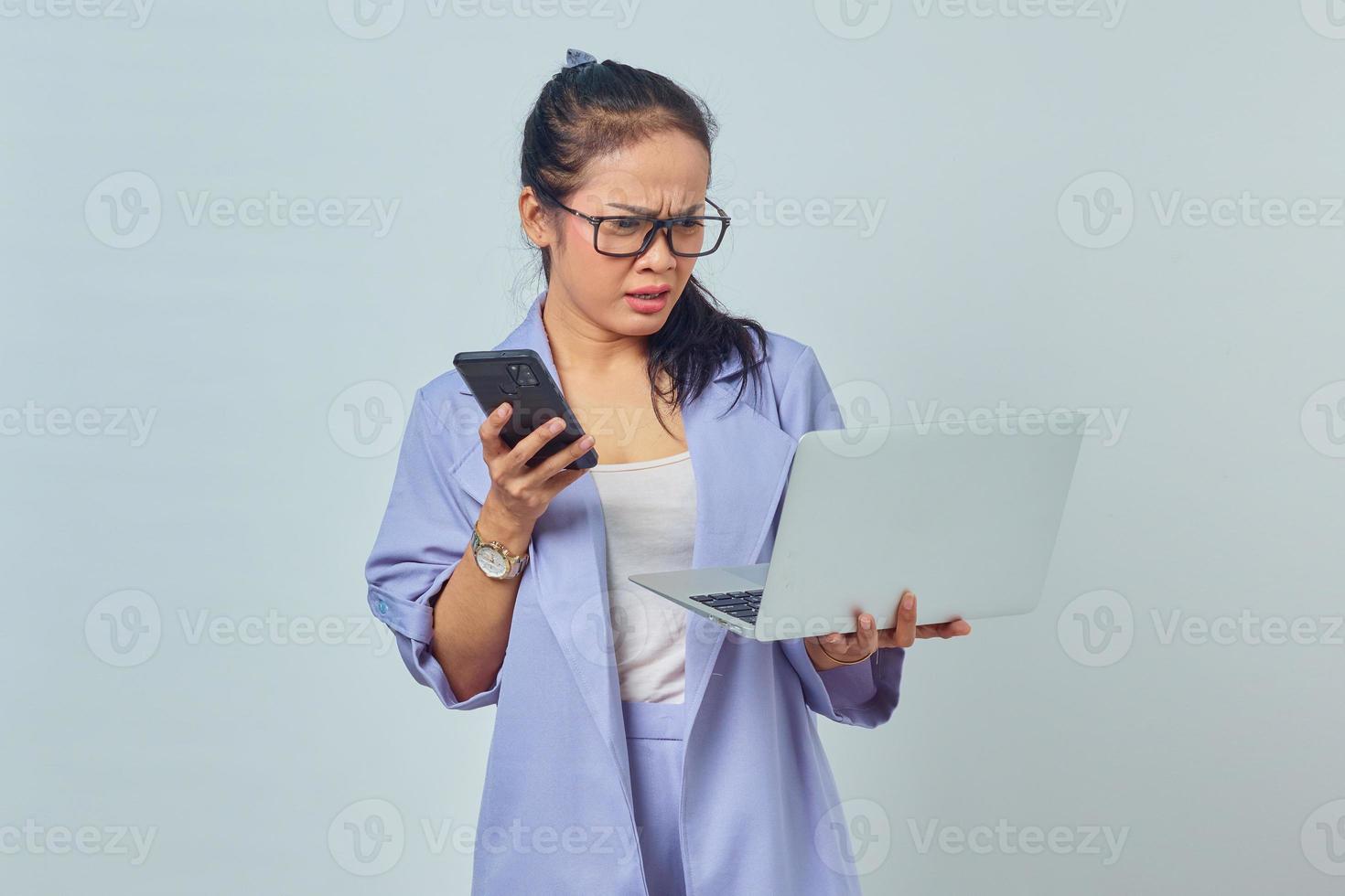 porträtt av förvånad ung asiatisk kvinna som håller smartphone och tittar på inkommande e-post på bärbar dator isolerad på vit bakgrund foto