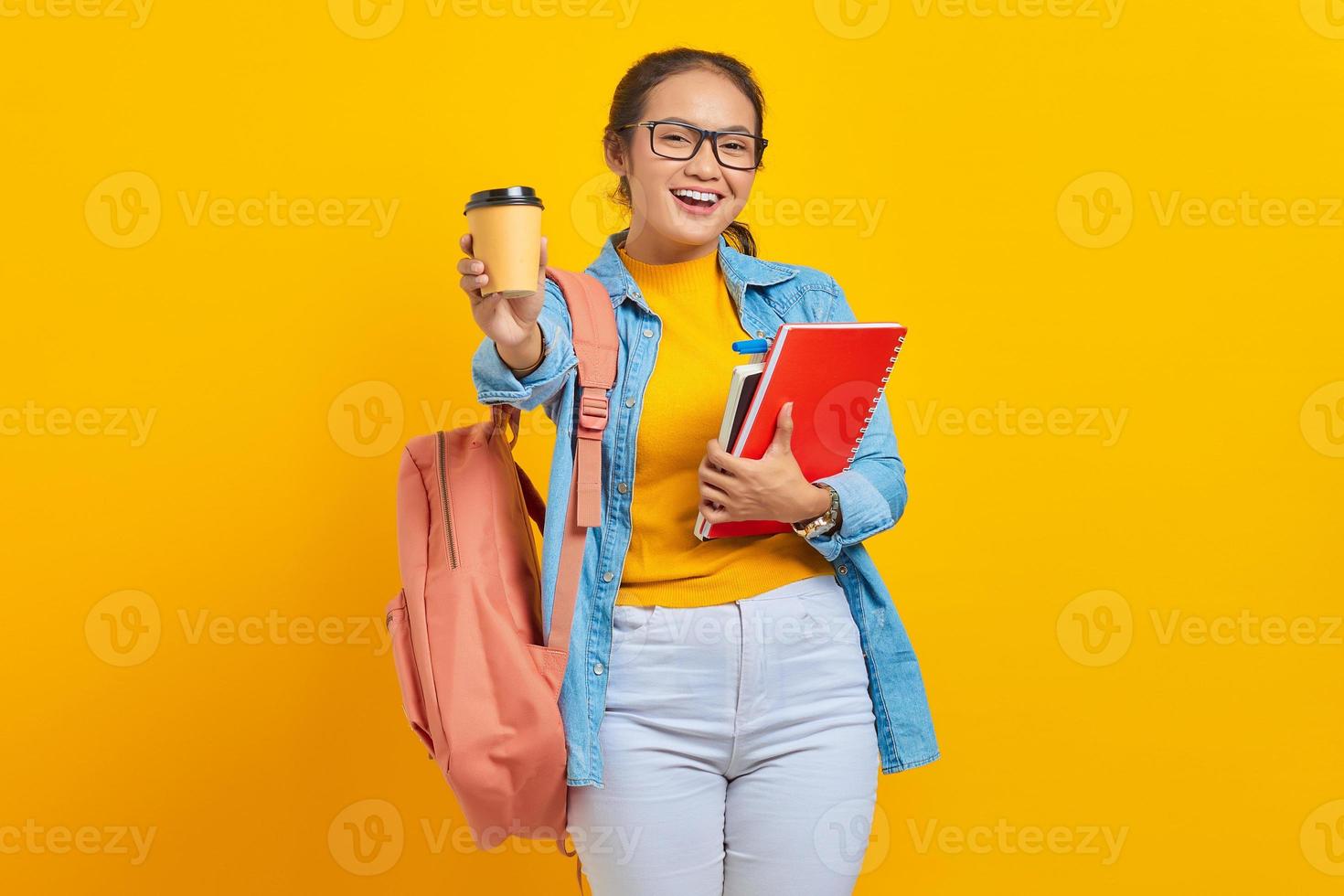 porträtt av glad ung asiatisk kvinna student i fritidskläder med ryggsäck som håller bok och visar kaffe take away tittar på kameran isolerad på gul bakgrund foto
