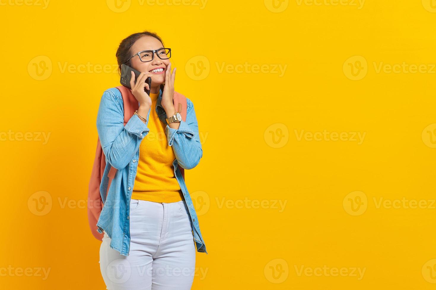 porträtt av upphetsad ung asiatisk kvinna student i fritidskläder med ryggsäck som håller smartphone och täcker mun med handflatorna isolerad på gul bakgrund. utbildning i college universitet koncept foto