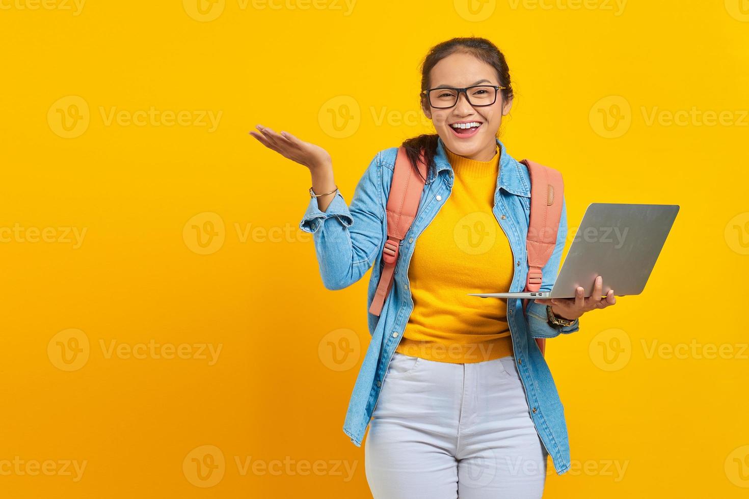 porträtt av leende ung asiatisk kvinna student i fritidskläder med ryggsäck visar kopia utrymme på handflatan och hålla laptop isolerad på gul bakgrund. utbildning i högskolekoncept foto