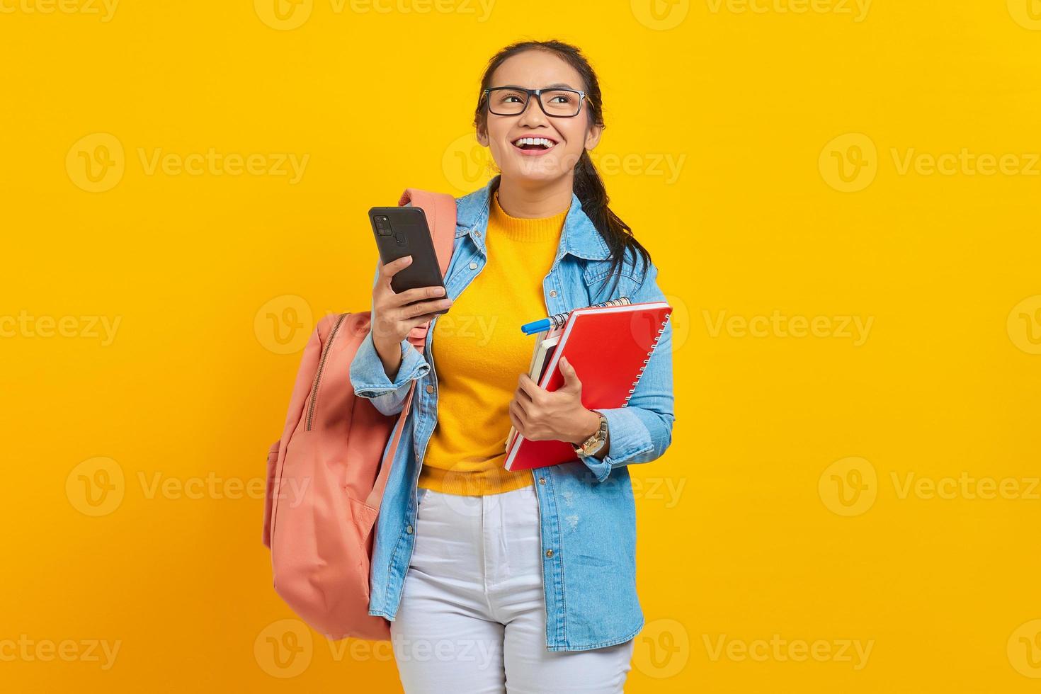 porträtt av leende ung asiatisk kvinna student i jeanskläder, glasögon med ryggsäck som håller mobiltelefon och böcker isolerad på gul bakgrund. utbildning i gymnasiekonceptet foto