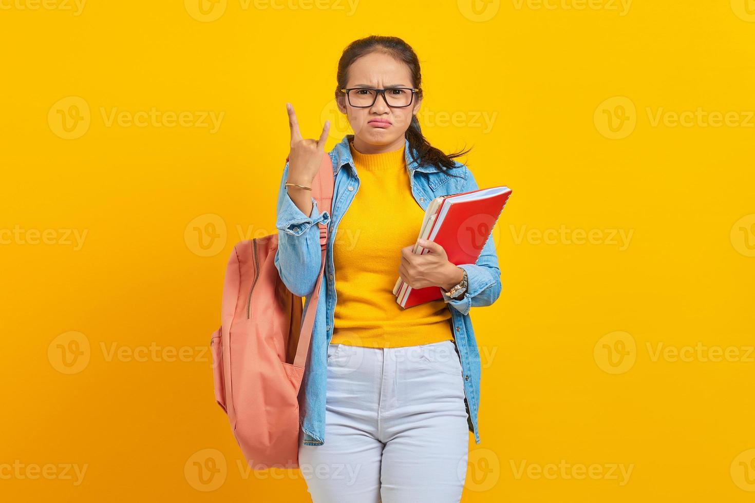 porträtt av vacker ung asiatisk student i jeanskläder med ryggsäck som håller anteckningsboken och skriker med galet uttryck som gör rocksymbol med händerna upp isolerade på gul bakgrund foto