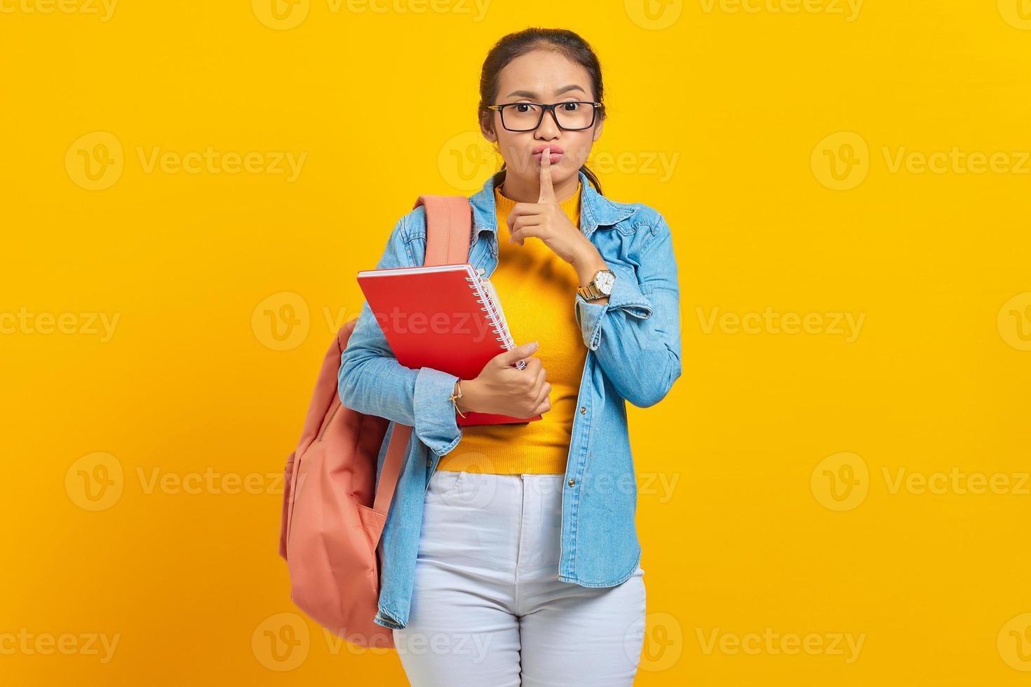 vacker ung kvinna student i jeanskläder med ryggsäck och hålla bok, fingrar som täcker läpparna, gör tyst gest på gul bakgrund. utbildning i gymnasiekonceptet foto