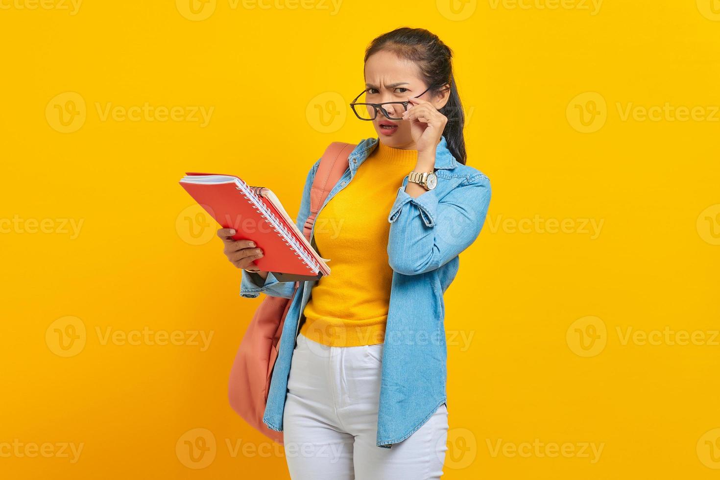 eftertänksam vacker ung asiatisk kvinna student i jeanskläder med ryggsäck som håller anteckningsboken och tittar på kameran isolerad på gul bakgrund. utbildning i gymnasiekonceptet foto