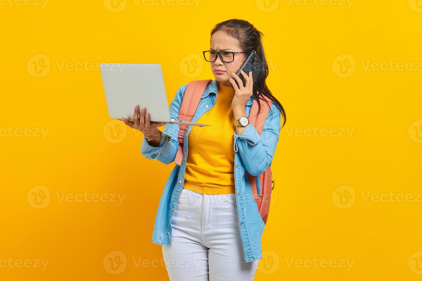 porträtt av förvirrad ung asiatisk kvinna student i fritidskläder med ryggsäck som pratar i mobiltelefon och håller bärbar dator isolerad på gul bakgrund. utbildning i college universitet koncept foto