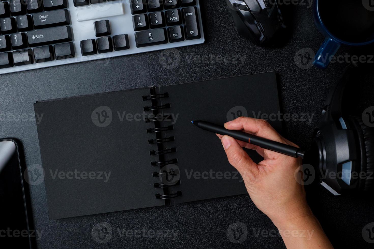 ovanifrån hand skriver på anteckningsboken arbetar på skrivbordet med tangentbord hörlurar på svart bordsbakgrund foto