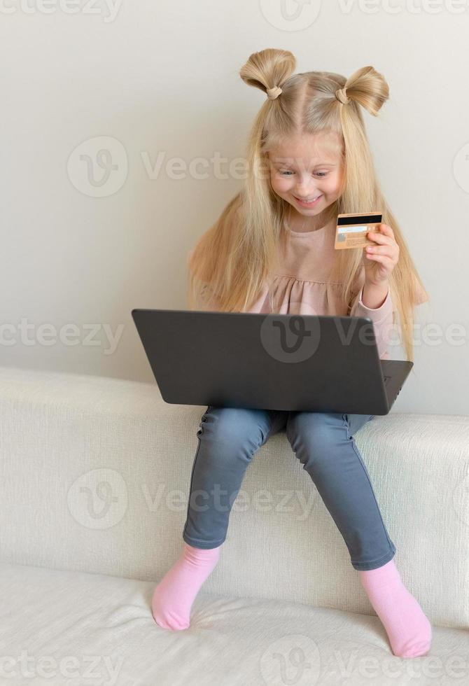 glad söt kaukasisk liten flicka håller kreditkort köpa online sitter på soffan foto