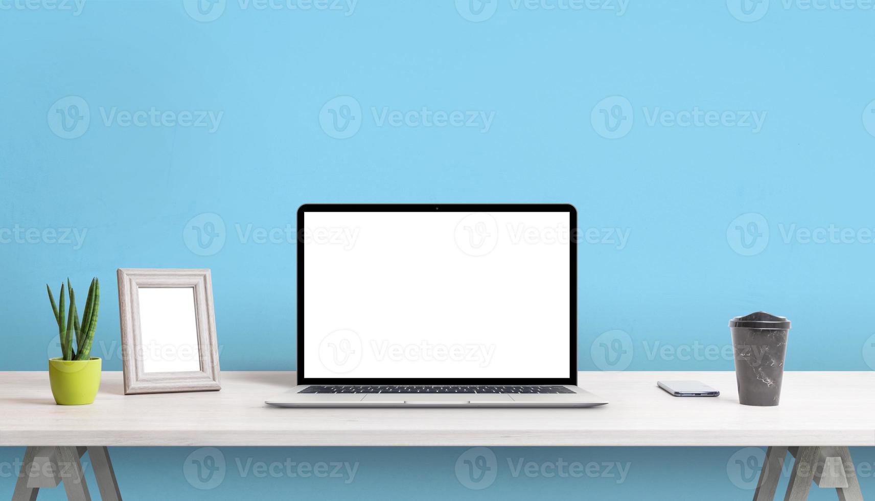 bärbar dator med tom skärm på skrivbordet. tavelram, växt, kaffe och telefon bredvid. blå vägg i bakgrunden foto