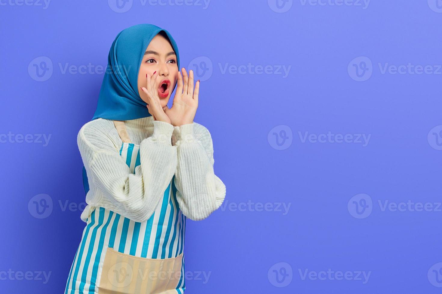 vacker ung asiatisk muslimsk kvinna i hijab och randigt förkläde, skrikande och ropade goda nyheter med händerna nära munnen isolerad på lila bakgrund. människor hemmafru muslimsk livsstilskoncept foto