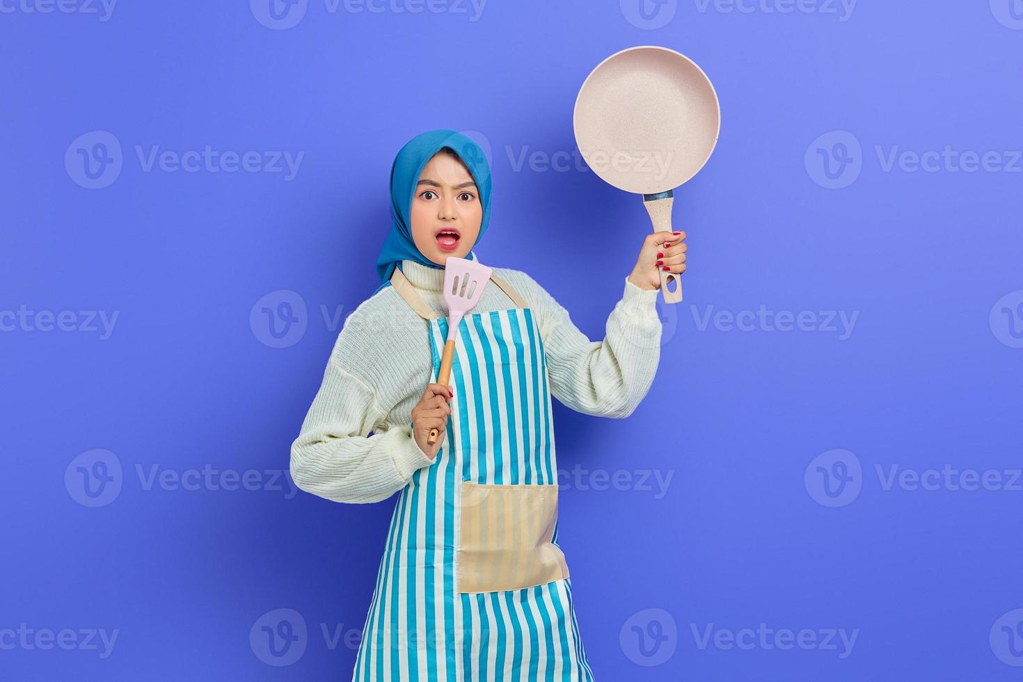 porträtt av förvånad ung asiatisk muslimsk kvinna hemmafru som bär förkläde håller stekpanna och täcker mun med spatel isolerad på lila bakgrund. hushållning koncept foto