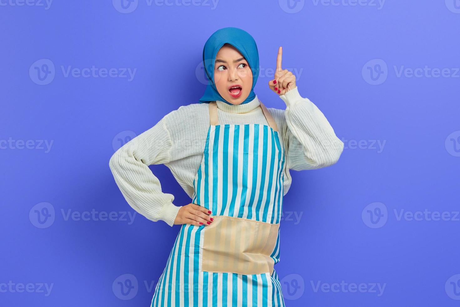 porträtt av chockad ung asiatisk muslimsk kvinna i hijab och randigt förkläde, öppen mun samtidigt som du pekar på kopieringsutrymme med fingret isolerat på lila bakgrund. människor hemmafru muslimsk livsstilskoncept foto