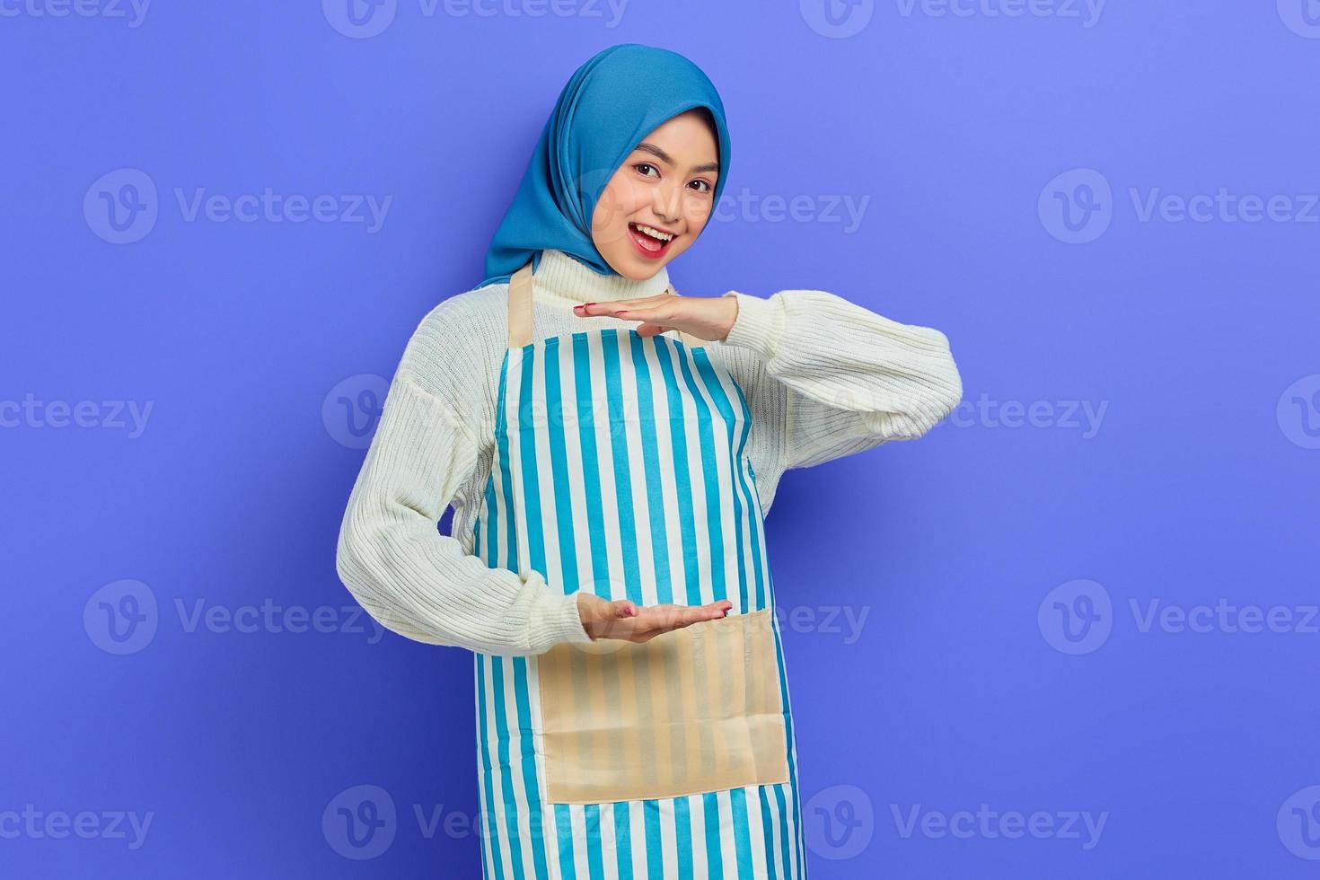 glad ung asiatisk muslimsk kvinna i hijab och randigt förkläde, visar produkt med palm, visar kopia utrymme för viss rabatt på lila bakgrund. människor hemmafru muslimsk livsstilskoncept foto