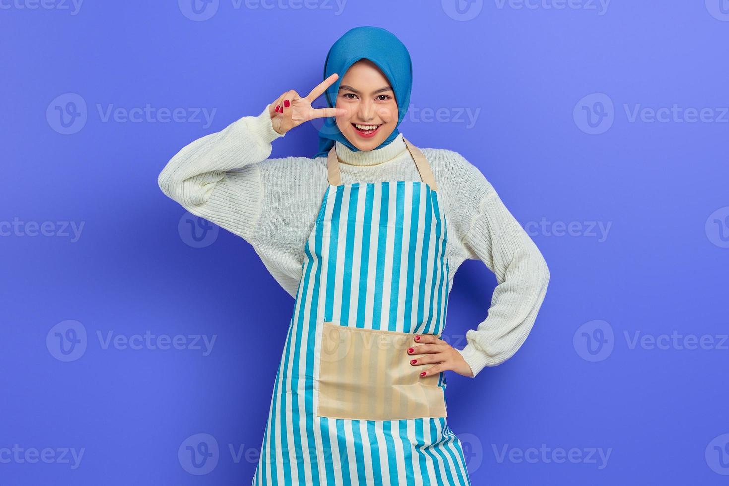 porträtt av glad ung hemmafru kvinna i hijab och randigt förkläde, täcker ögat med finger, seger v-tecken gest isolerad på lila bakgrund. människor hemmafru muslimsk livsstilskoncept foto