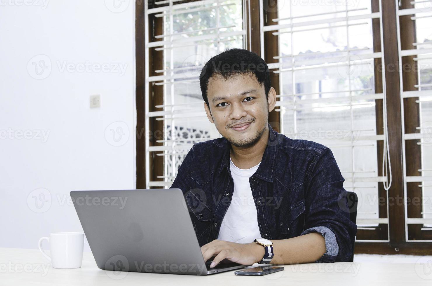 ung asiatisk man känner sig glad och ler när arbete laptop på bordet. indonesisk man klädd i blå skjorta. foto