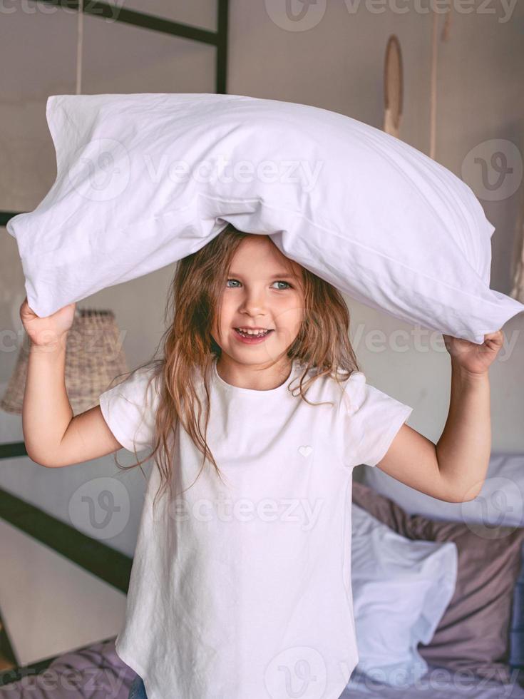 bedårande glad liten flicka i vit t-shirt slåss kudde hemma inomhus. mode, roligt, stil, barndom, känslor, uppväxt koncept foto