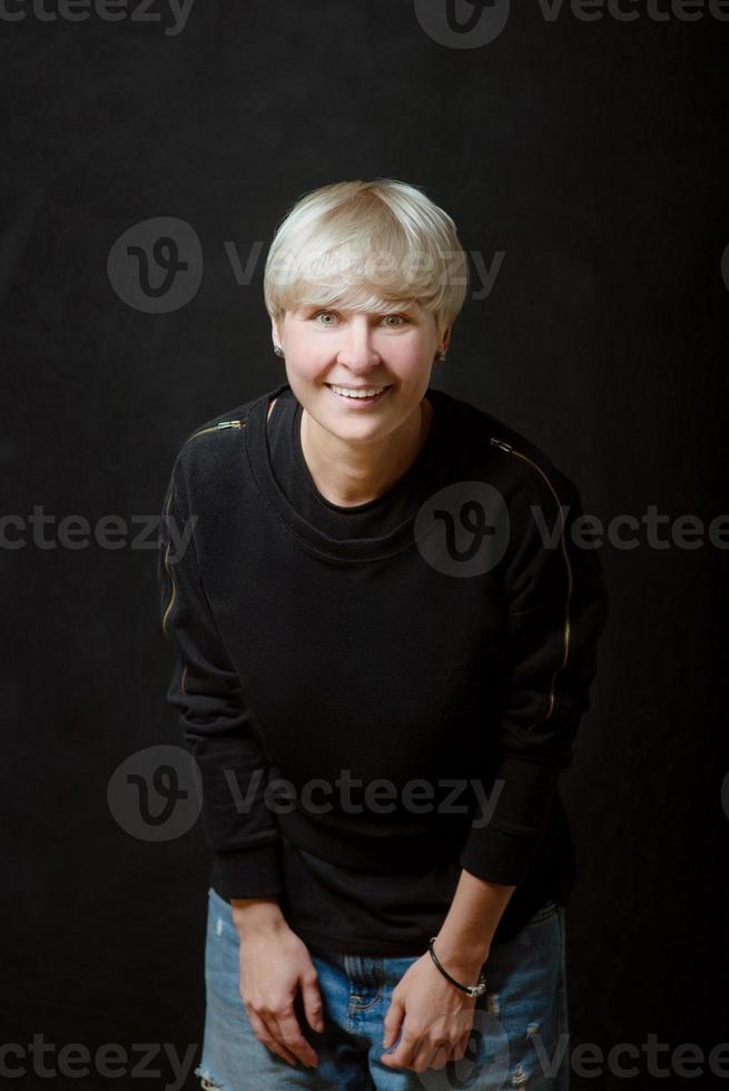 porträtt av snygg blond kort hår ung kvinna i svart tröja och jeans på mörk bakgrund foto