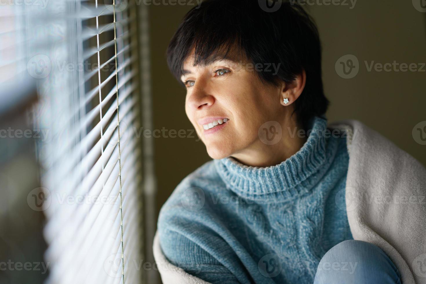 glad medelålders kvinna ler medan du tittar ut genom fönstret. foto
