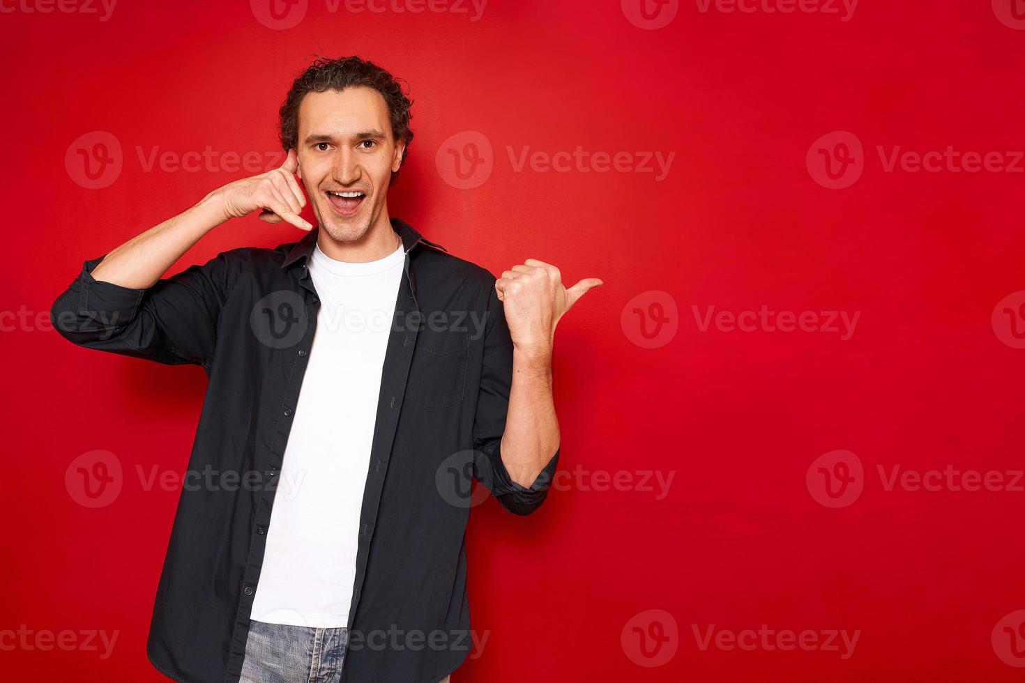 handtecken ring mig. porträtt av ung leende man pekar med tummen på ledigt tomt utrymme för en reklamtext och föreställer en telefonlur med handen. isolerad på en röd studio bakgrund foto