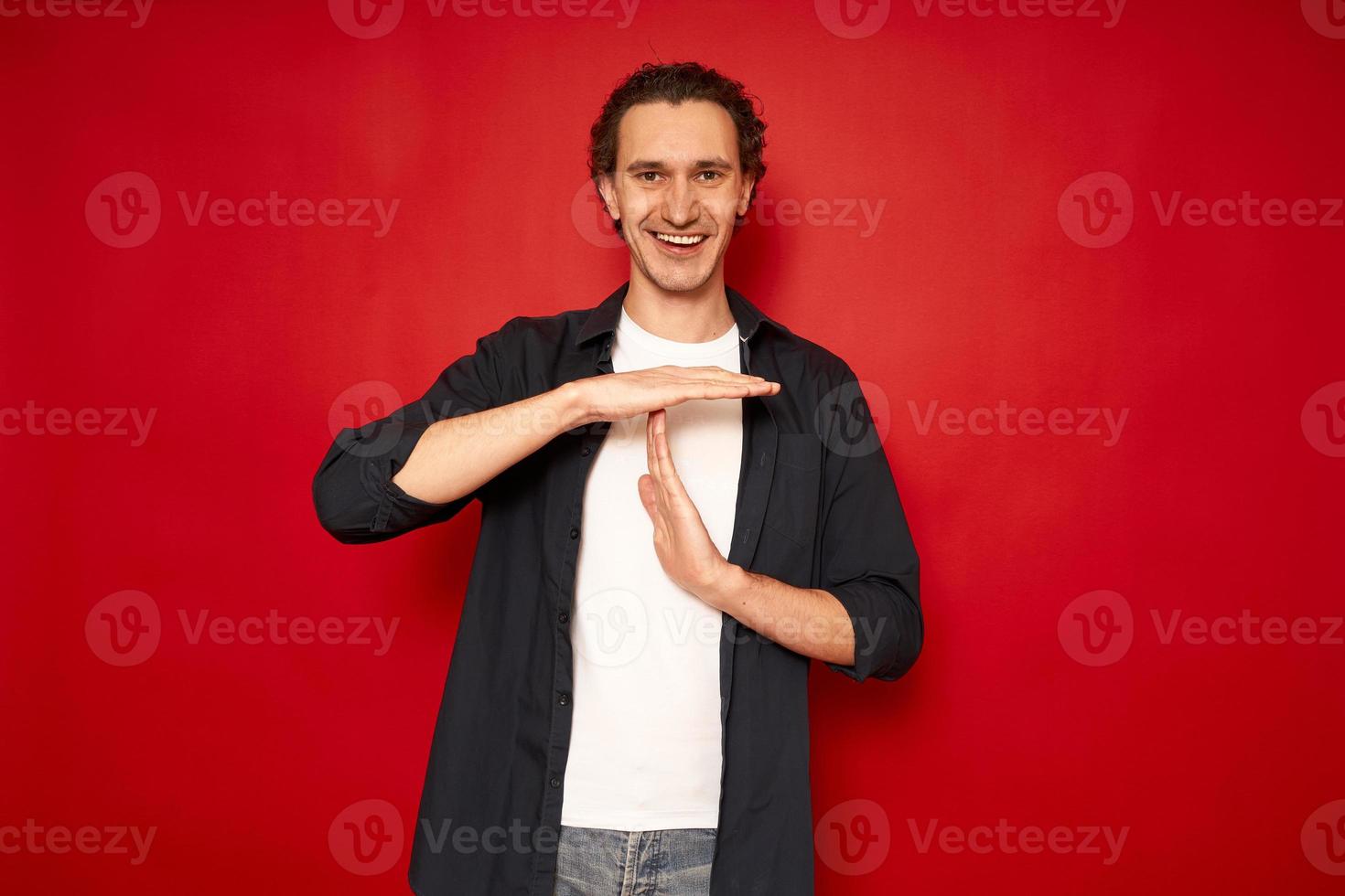 studiobild av glada leende man fritidskläder visar timeout gest behöver stopp ber tid för vila efter hårt arbete visar paus hand tecken. isolerad av röd bakgrund kroppsspråk människor koncept foto