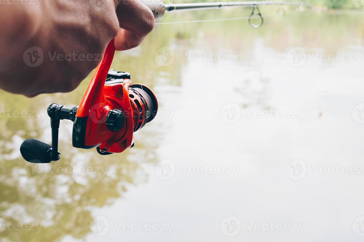 närbild av spinning med fiskerullen i handen, fiskekrok på linan med betet i vänster hand mot bakgrund av vattnet. foto