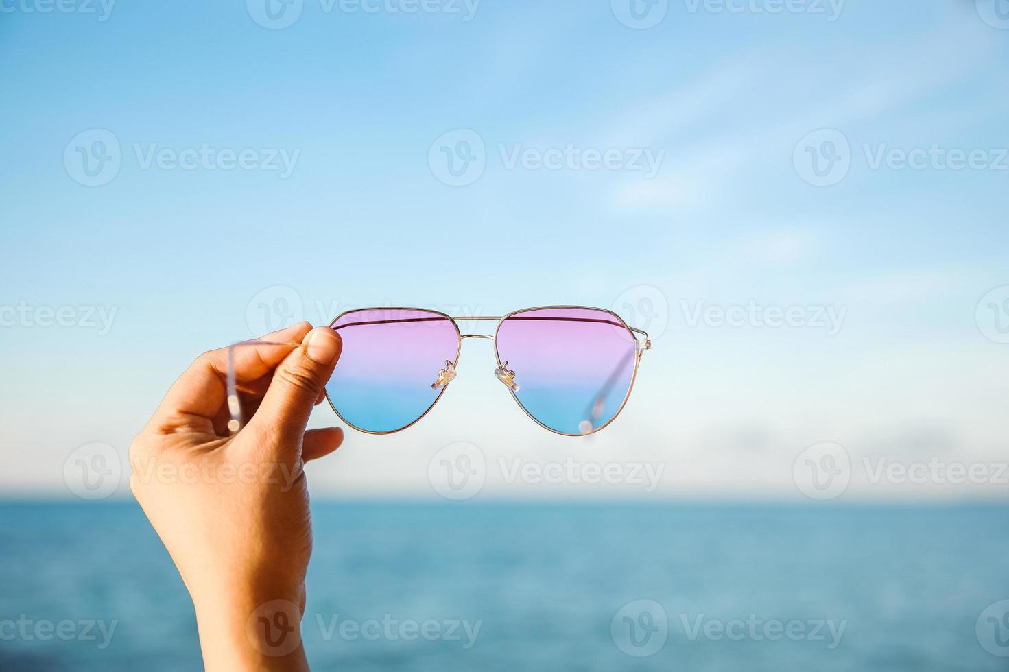 selektiv inriktning av hand som håller solglasögon med havet och blå himmel bakgrund foto