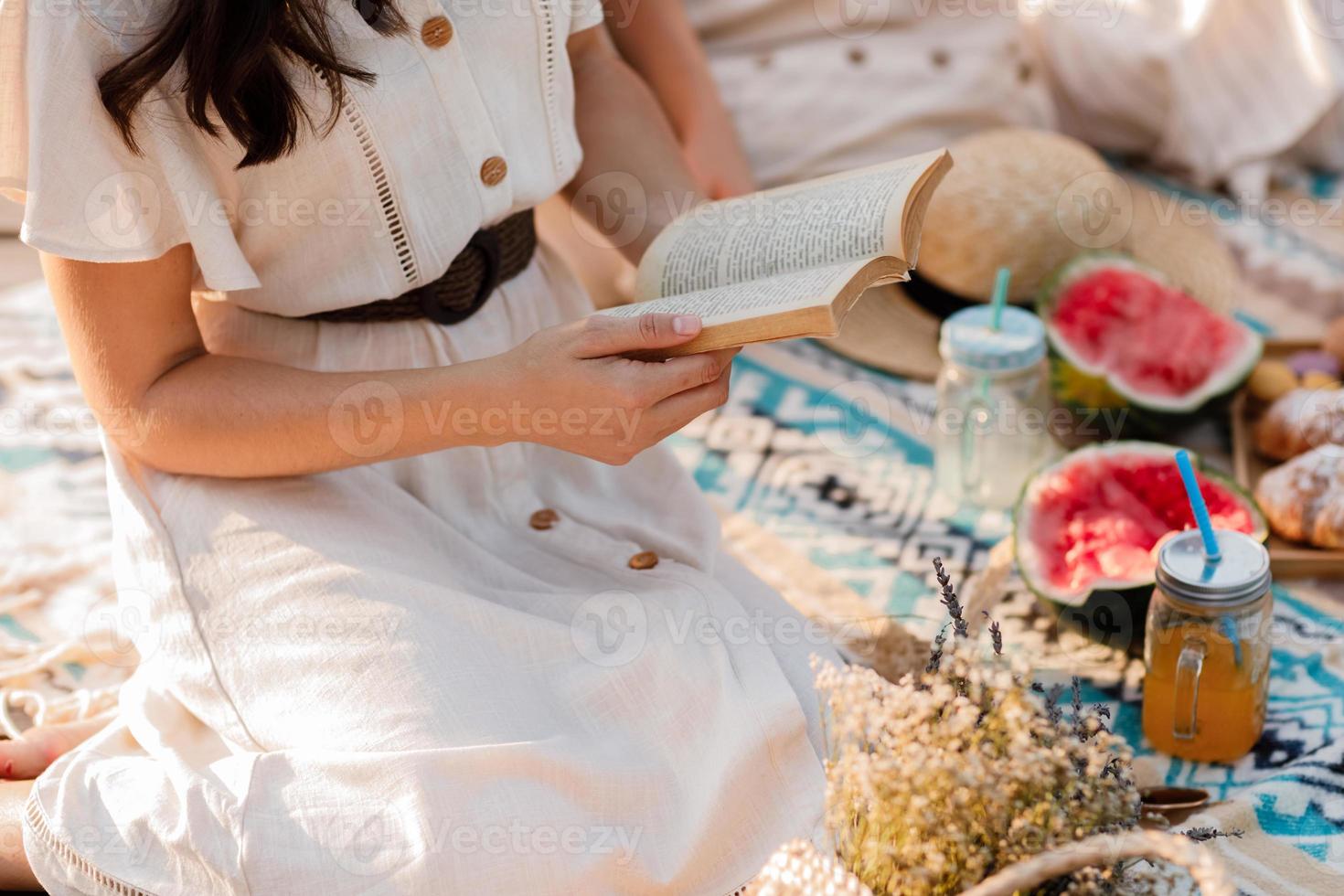 beskuret foto av ung kvinna i klänning som läser bok medan du njuter av picknick i sommarparken. sitter på filt. croissanter, vattenmelon och kall sommardrink