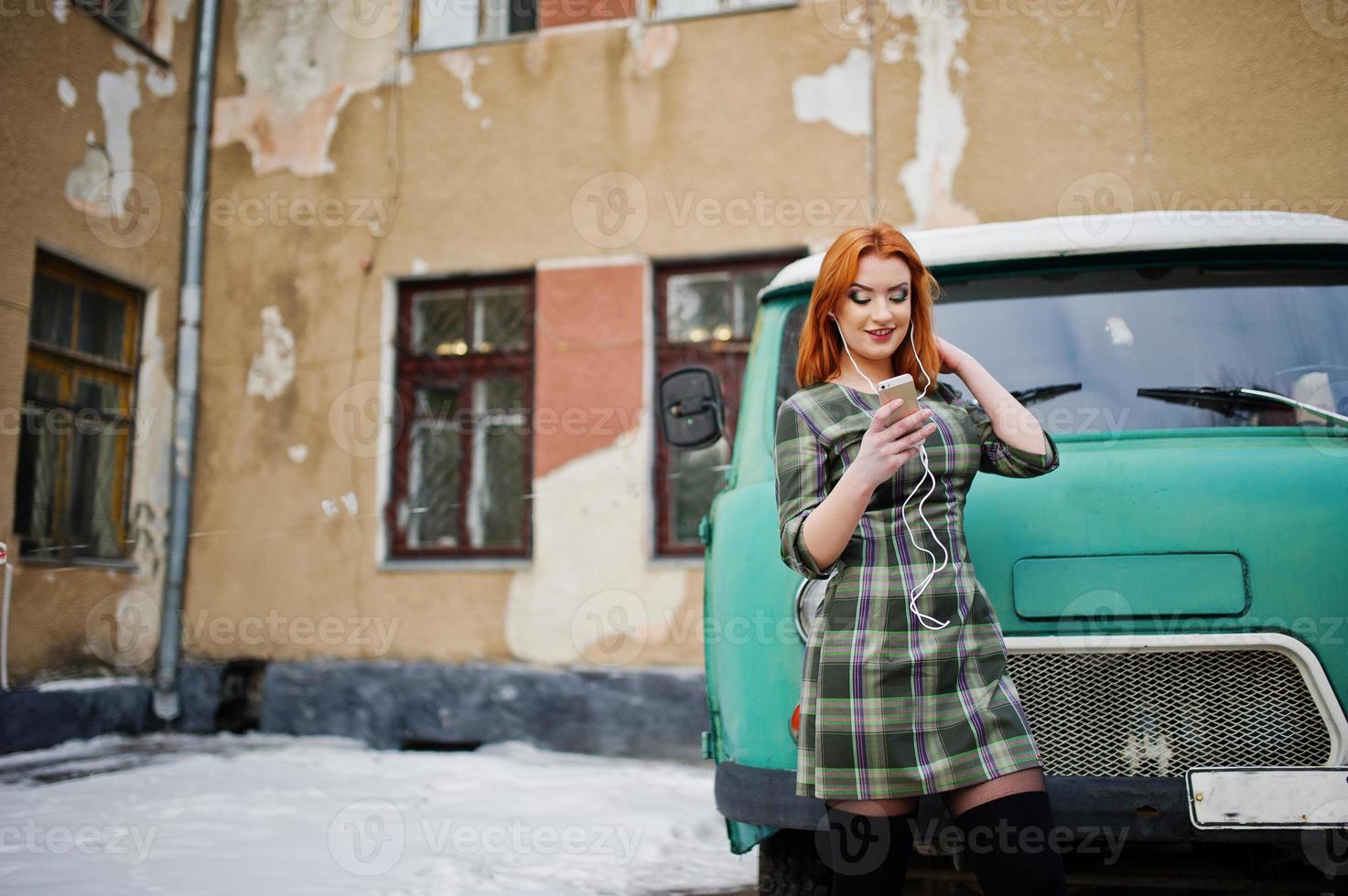 ung rödhårig flicka dam med mobiltelefon och hörlurar, bär på rutig klänning bakgrund gammal vintage turkos minibuss. foto