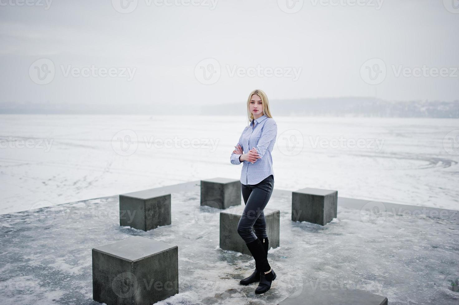 snygg rik ung blond tjej på blus och läderbyxor med rosa smartphone på sidan bakgrund stenkuber mot frusen sjö på vinterdagen. foto